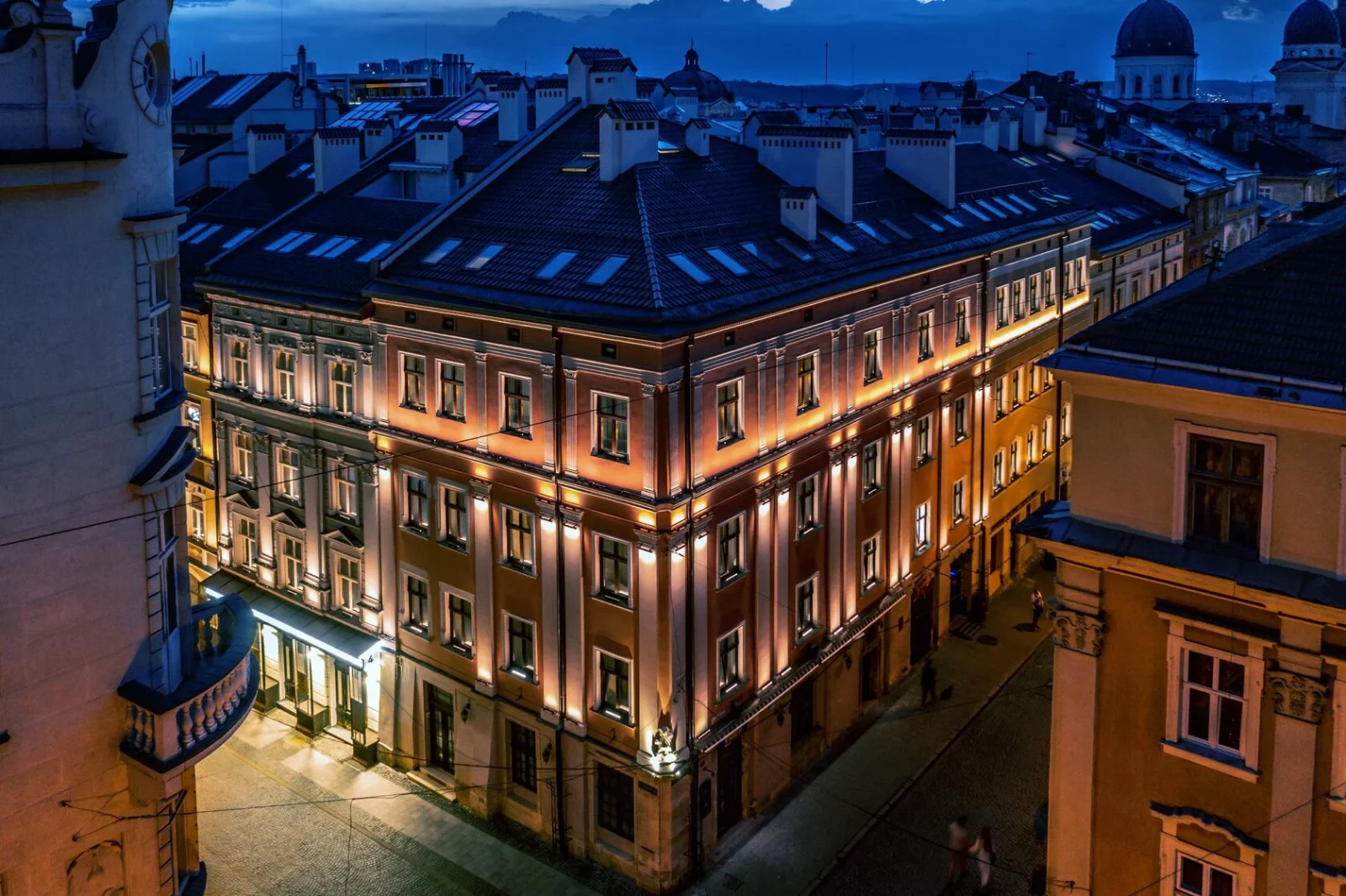 The city at dusk from above: a floodlit corner building with mansard roofs