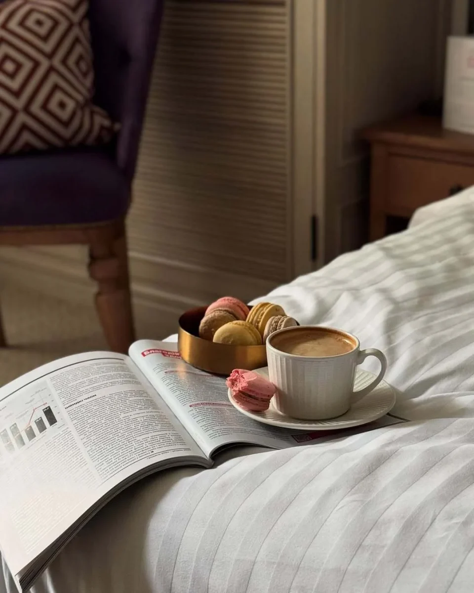 A cup of coffee and macarons beside an open magazine on white bedding